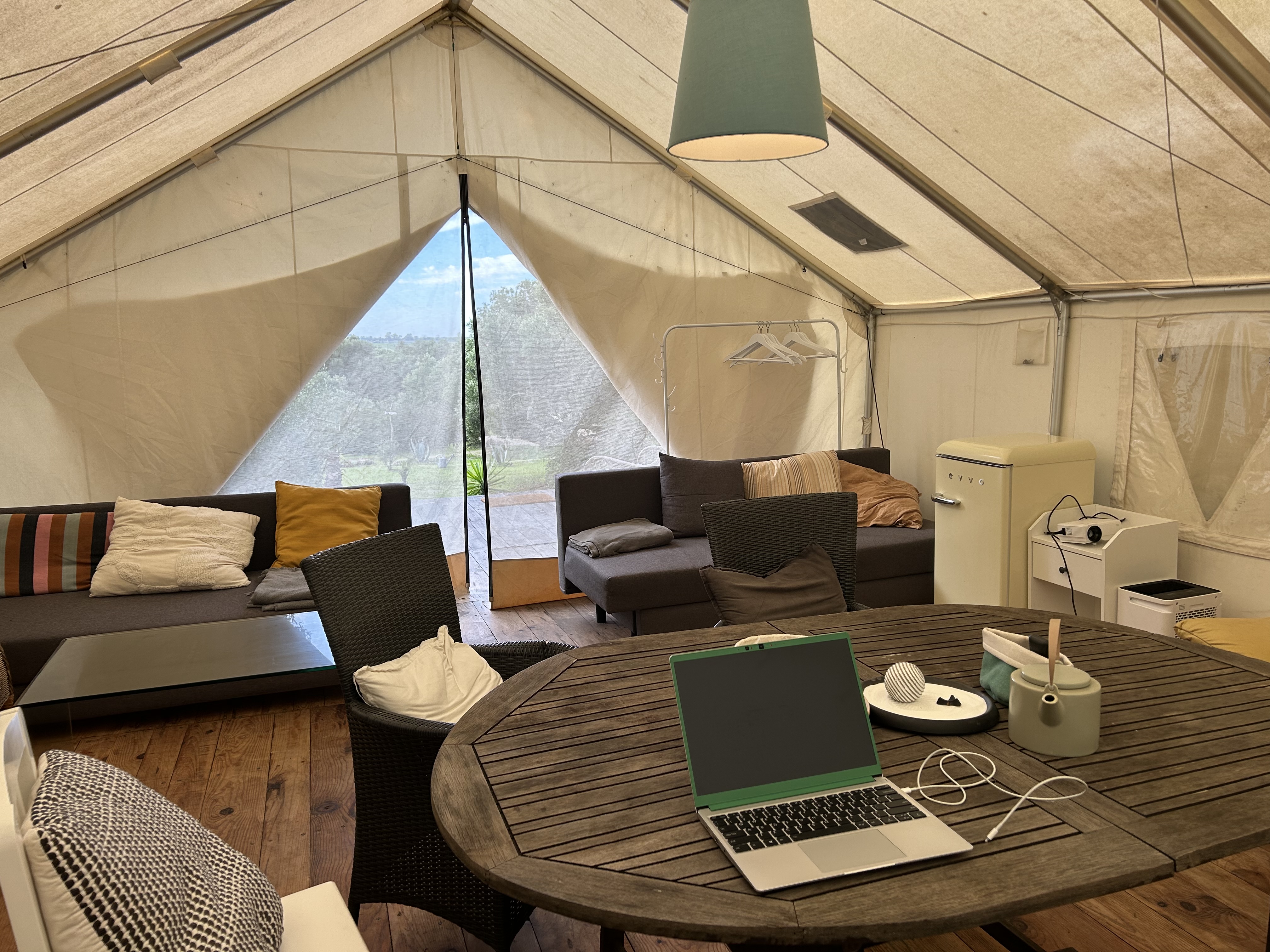 Zdjęcie wnętrza dużego namiotu z biurkiem i leżącym na nim laptopem, kanapami, fotelami i lodówką.